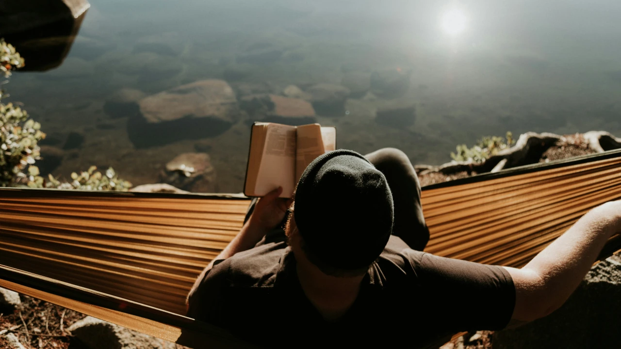 Man relaxing in a hammock by the lake while reading a book at sunset